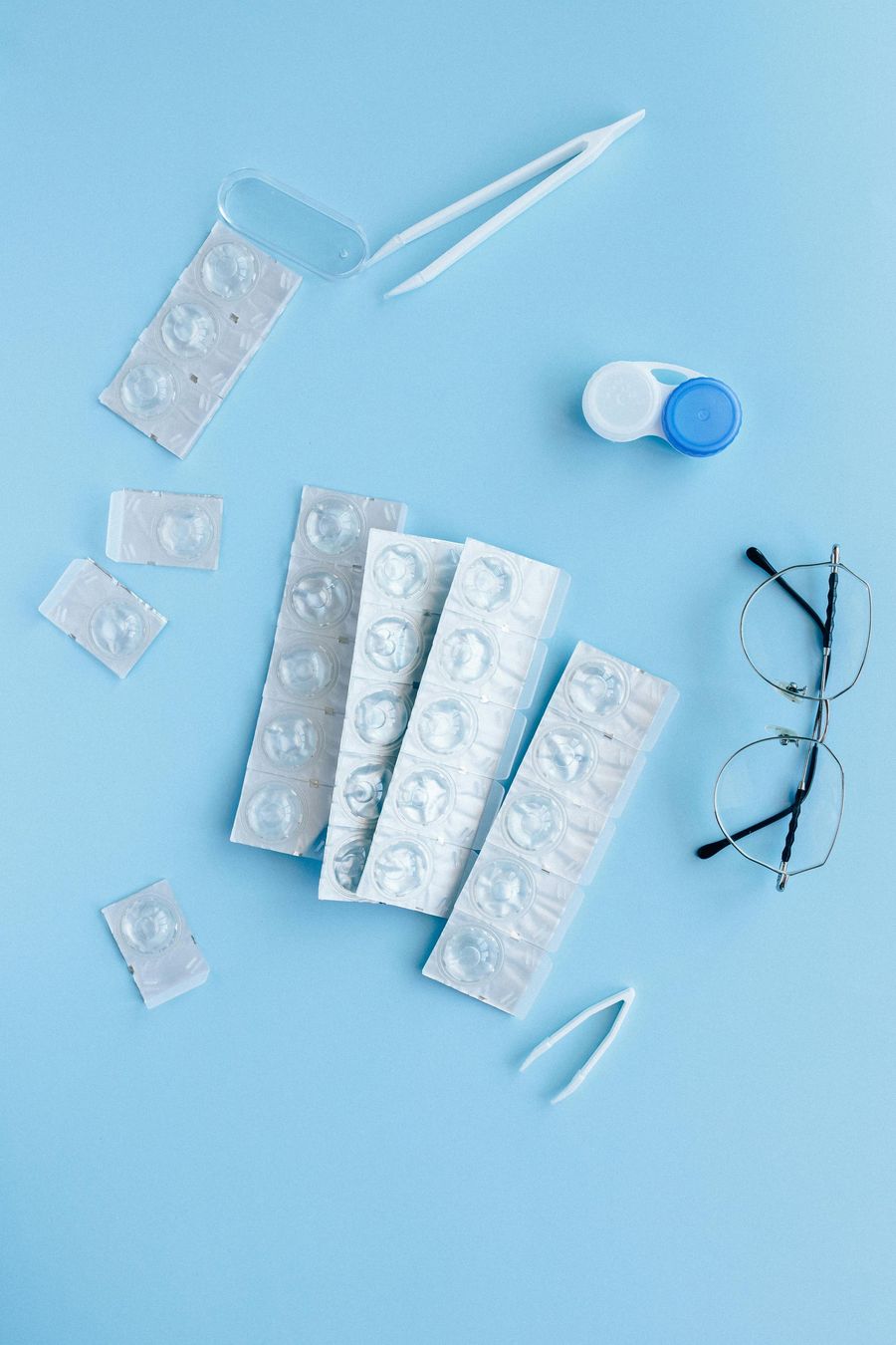 Contact lenses, tweezers, lens case, and eyeglasses on a light blue background