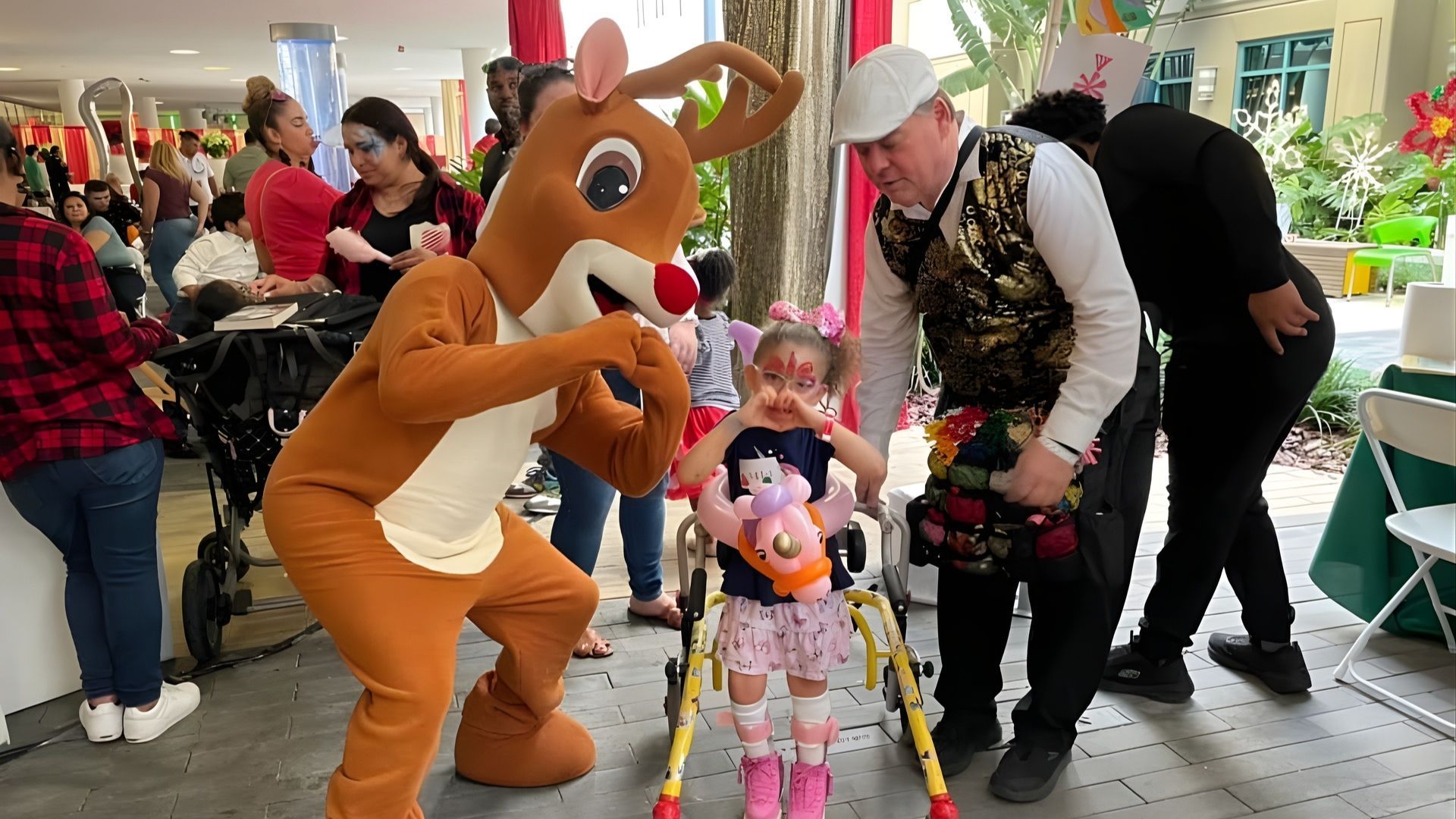 Rudolph the Red-Nosed Reindeer with a young child using a walker, surrounded by people at an outdoor event.