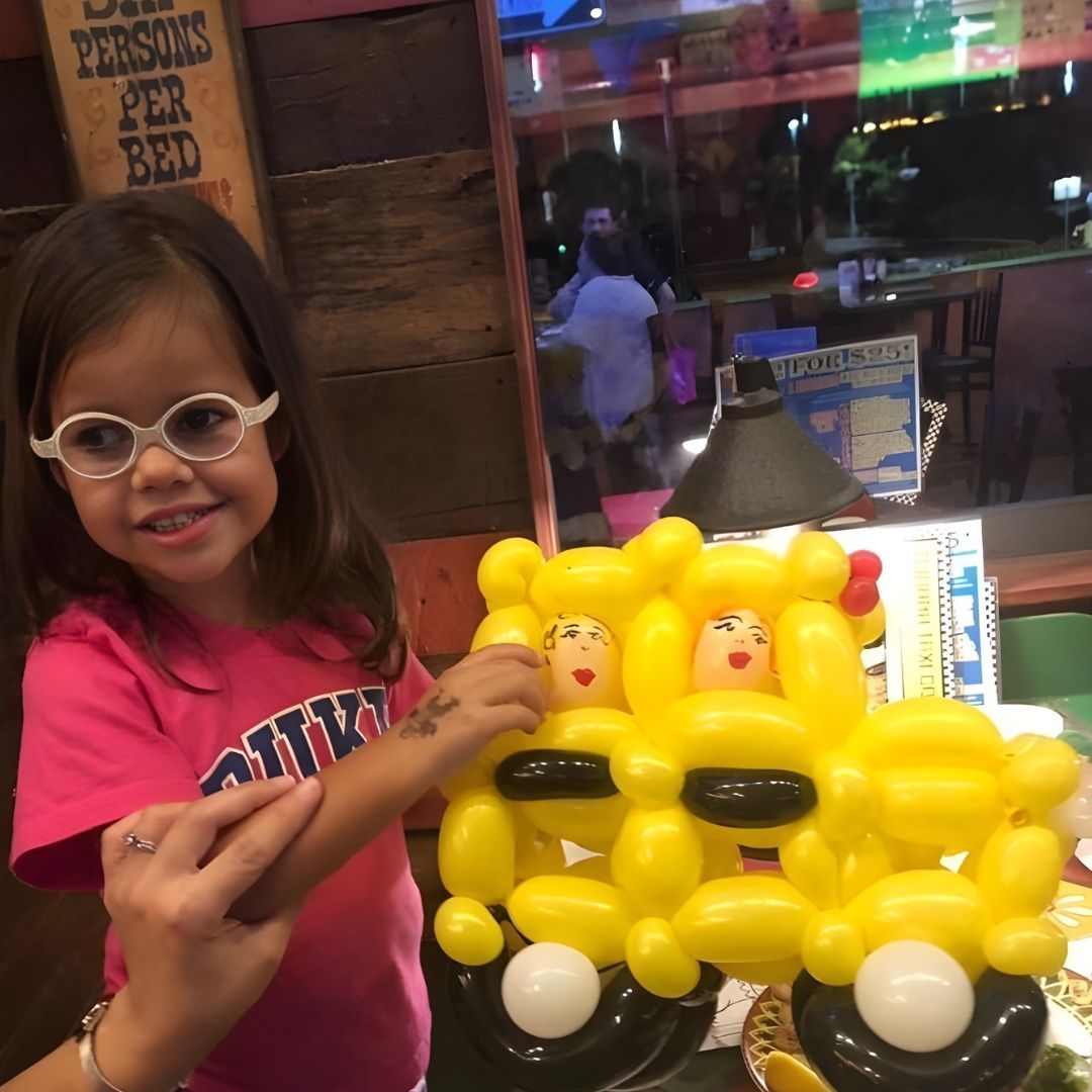 Young child with glasses smiles, holding a yellow balloon bus with two balloon doll passengers, inside a restaurant.