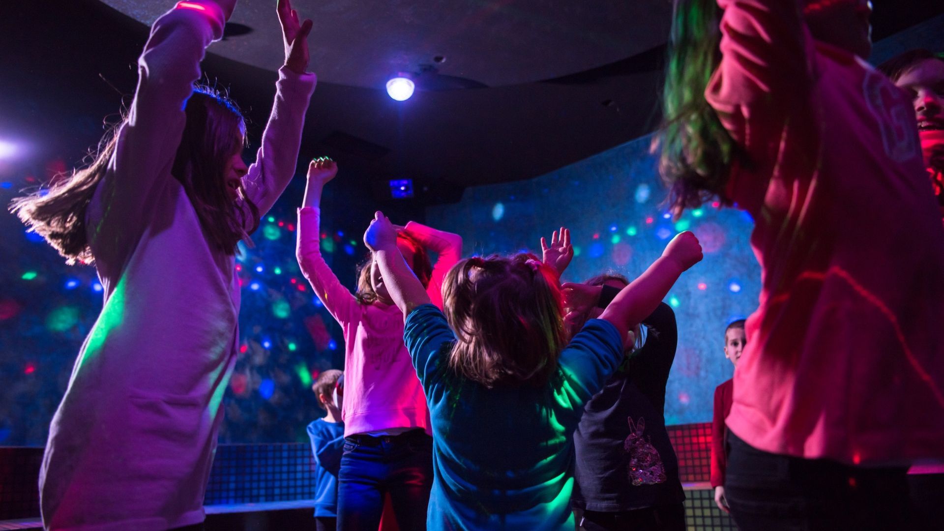 Children dancing in a brightly lit room with colorful lights, arms raised.