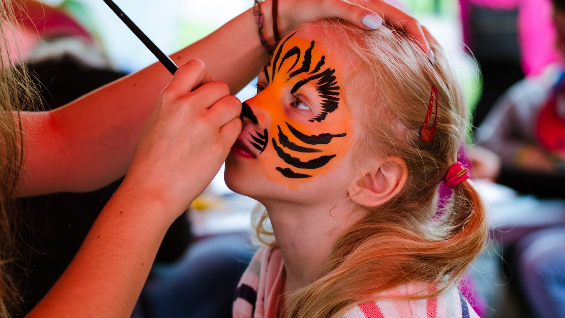 Girl getting tiger face paint applied at an outdoor event, orange and black colors.