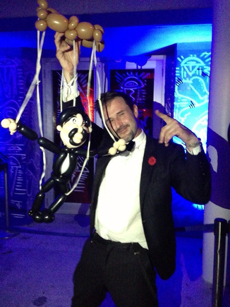 Magician in purple vest performing on stage with raised hands, audience in foreground.