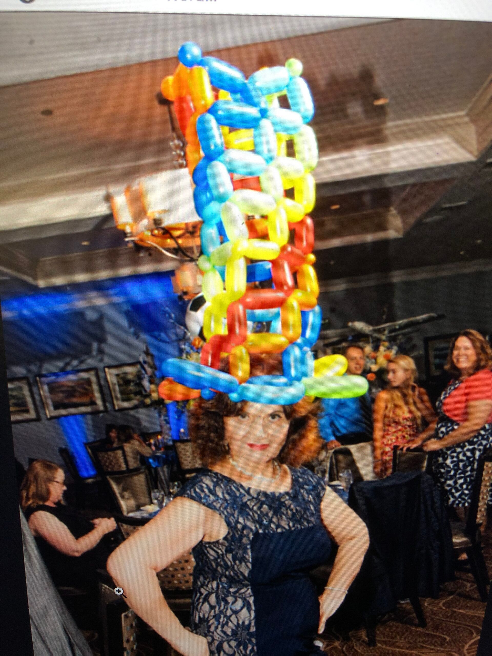 Woman wearing a colorful balloon hat, arms at sides, smiles at the camera, inside a room with other people.