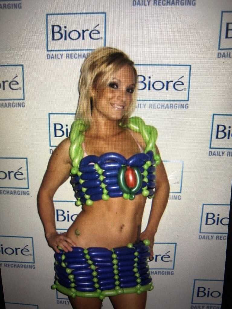 Woman wearing a colorful balloon hat, arms at sides, smiles at the camera, inside a room with other people.