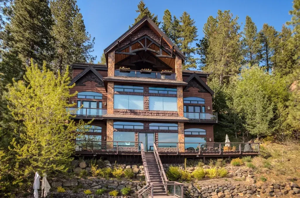 Large wooden lakeside house with multiple levels and large windows, surrounded by trees.