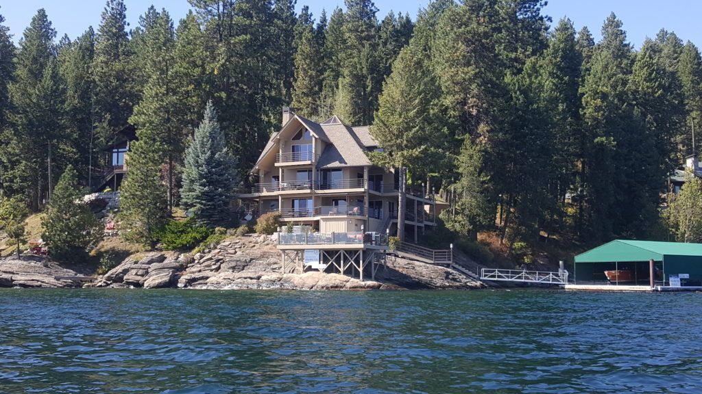 Lakeside multi-story house with decks, dock, and boat, surrounded by trees under a blue sky.