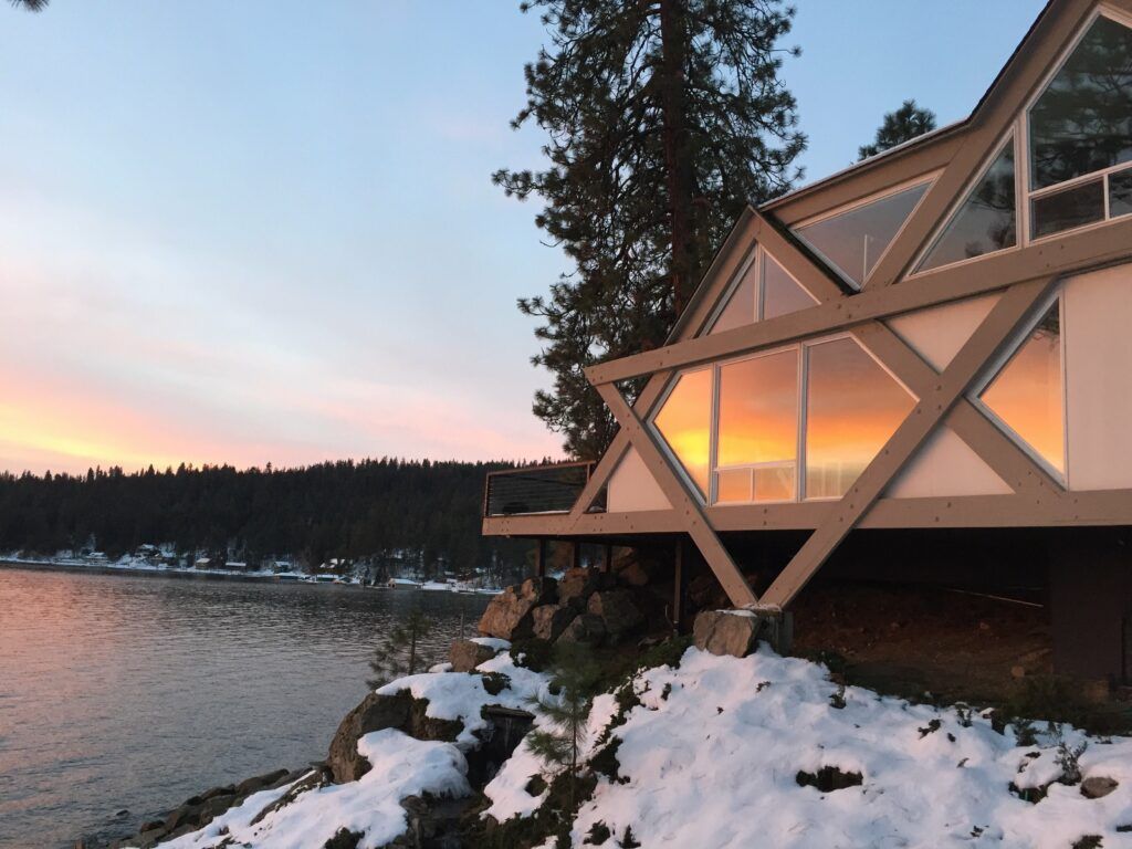 A-frame cabin overlooking a snowy lake at sunset; orange and pink hues reflect in the windows.