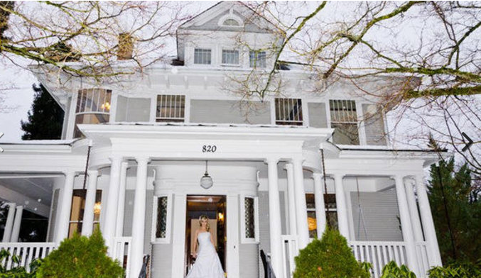Bride in white dress at the entrance of a white Victorian house, with porch and bare tree branches.