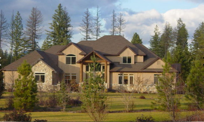 Beige two-story house with brown roof, stone accents, surrounded by trees and a green lawn under a cloudy sky.
