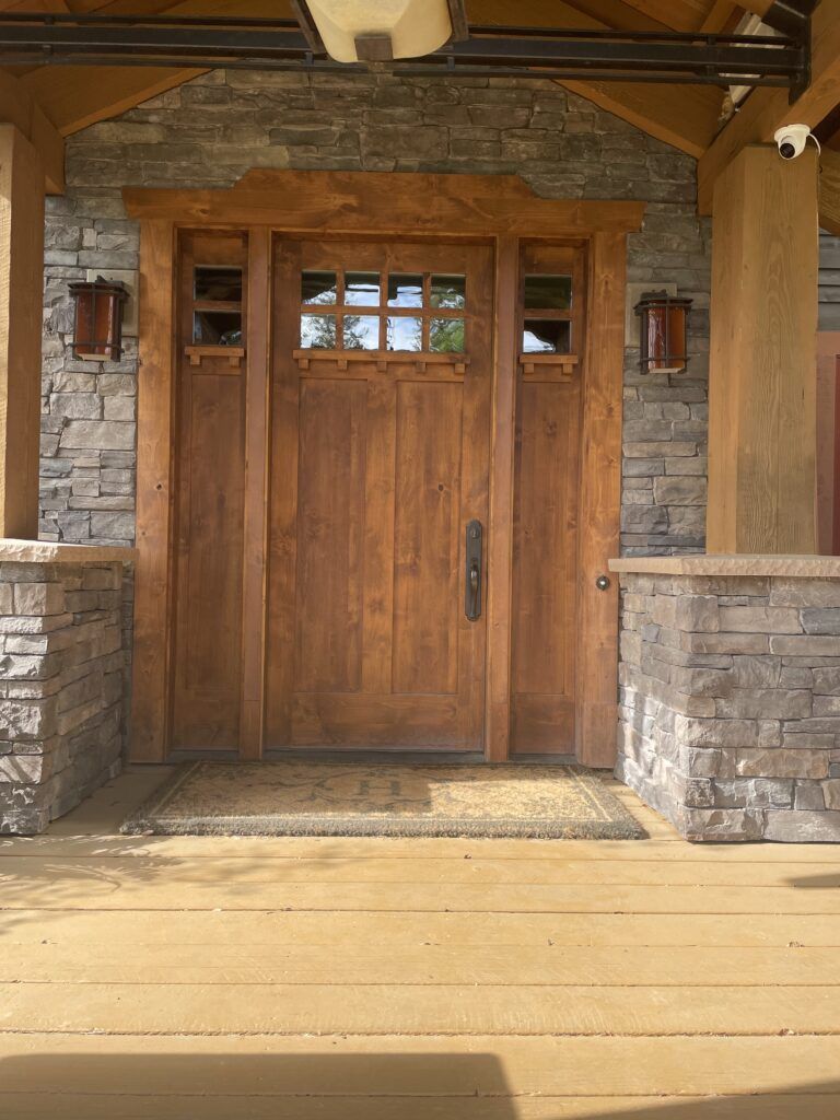Wooden door with sidelights and window, framed by stone and wood, with porch lights and a doormat.