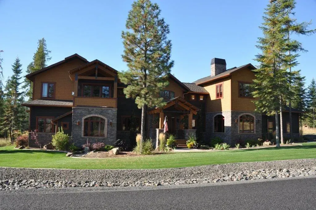 Large two-story house with stone and brown siding, trees, green lawn, and blue sky.