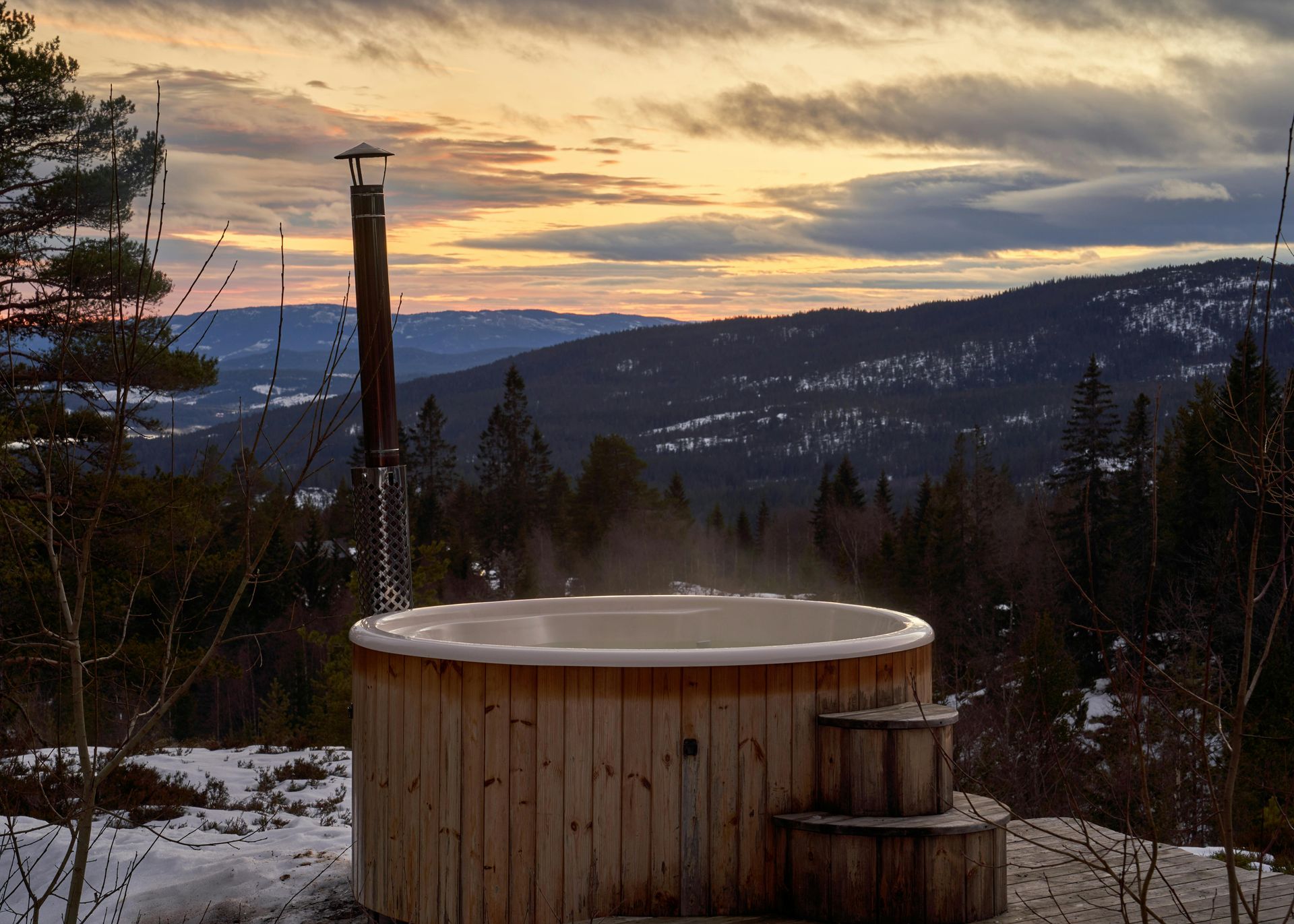 Wooden hot tub on a snowy hillside, smoke rising against a sunset over mountains and trees.