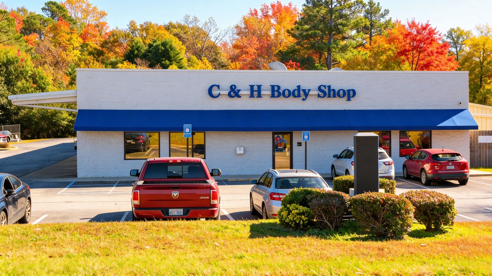 C & H Body Shop, a white building with blue awning, red pickup truck parked in front, autumn trees in the background.
