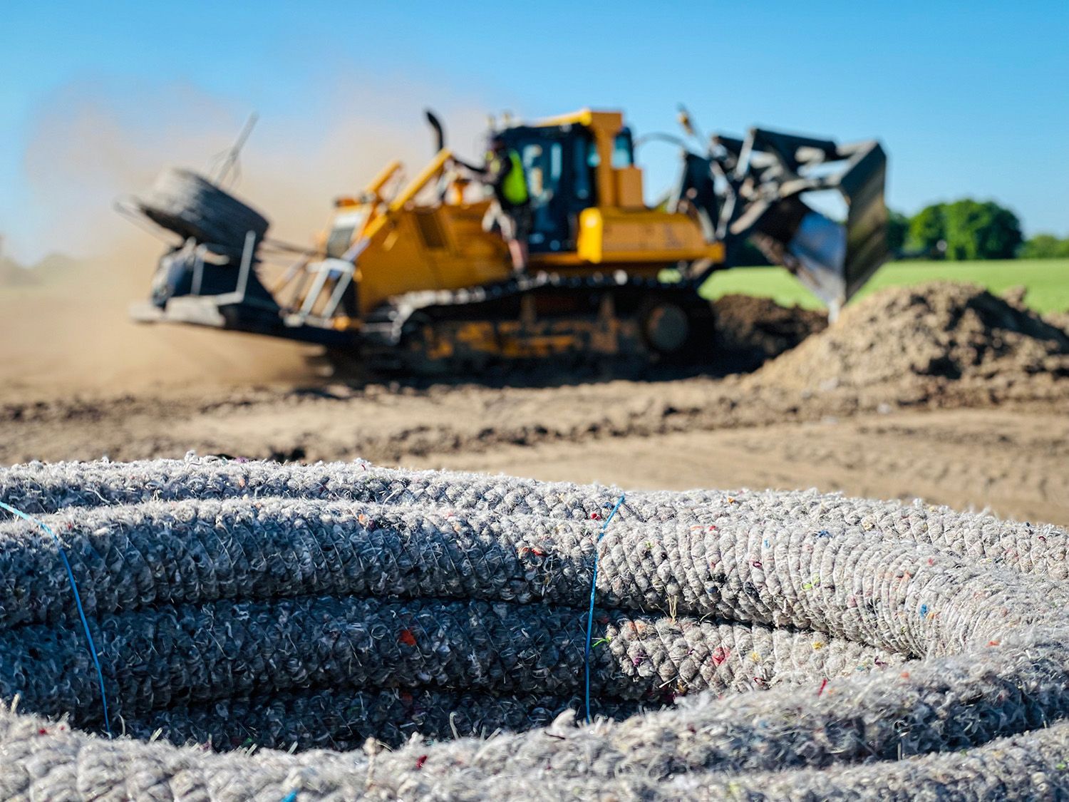 Op de voorgrond een opgerolde grijze buis voor landschapsinrichting, met op de achtergrond een gele bulldozer aan het werk.