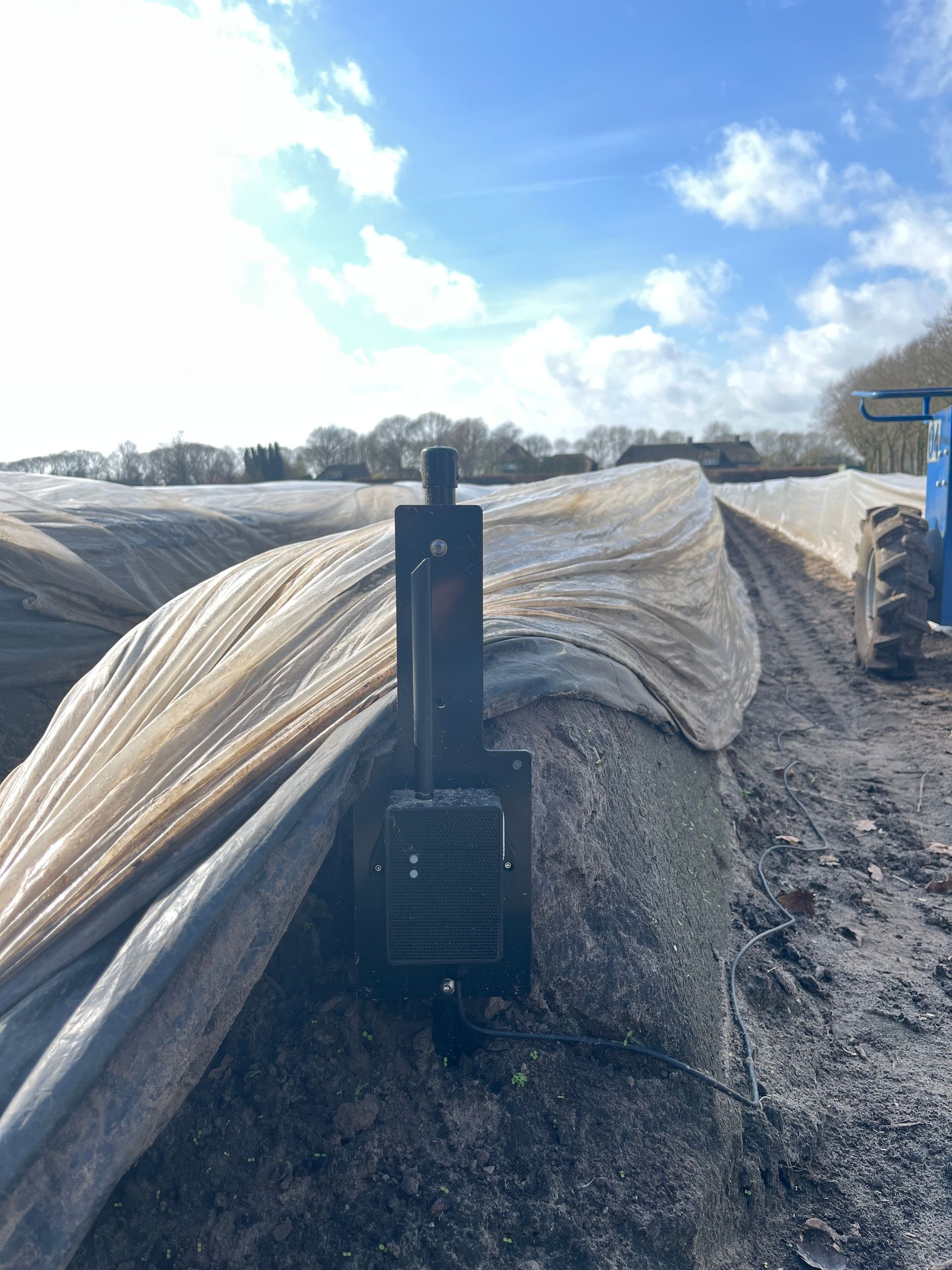 Een zwarte, verticale paal met een sensor staat in een landbouwveld, naast lange rijen grond die bedekt zijn met plastic folie.
