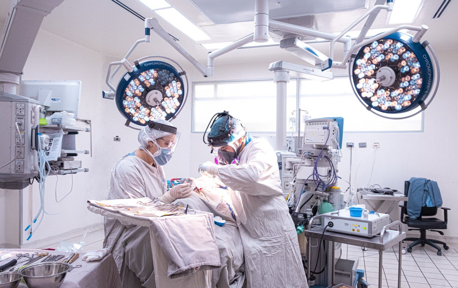 Dos cirujanos en un quirófano; uno de ellos operando a un paciente, iluminados por luces superiores.