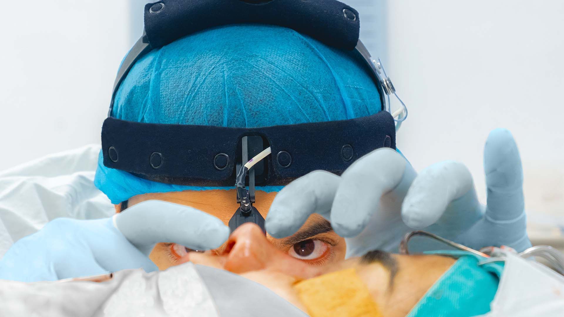 Cirujano con gorra azul, guantes y linterna frontal operando el rostro de un paciente en un ambiente estéril.