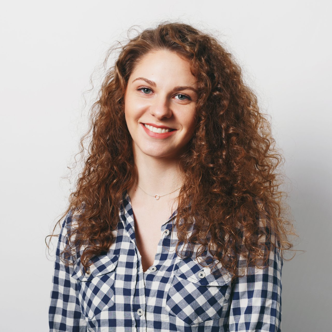 Mujer con cabello castaño rizado, sonriente, vistiendo una camisa a cuadros azul y blanca sobre un fondo blanco.