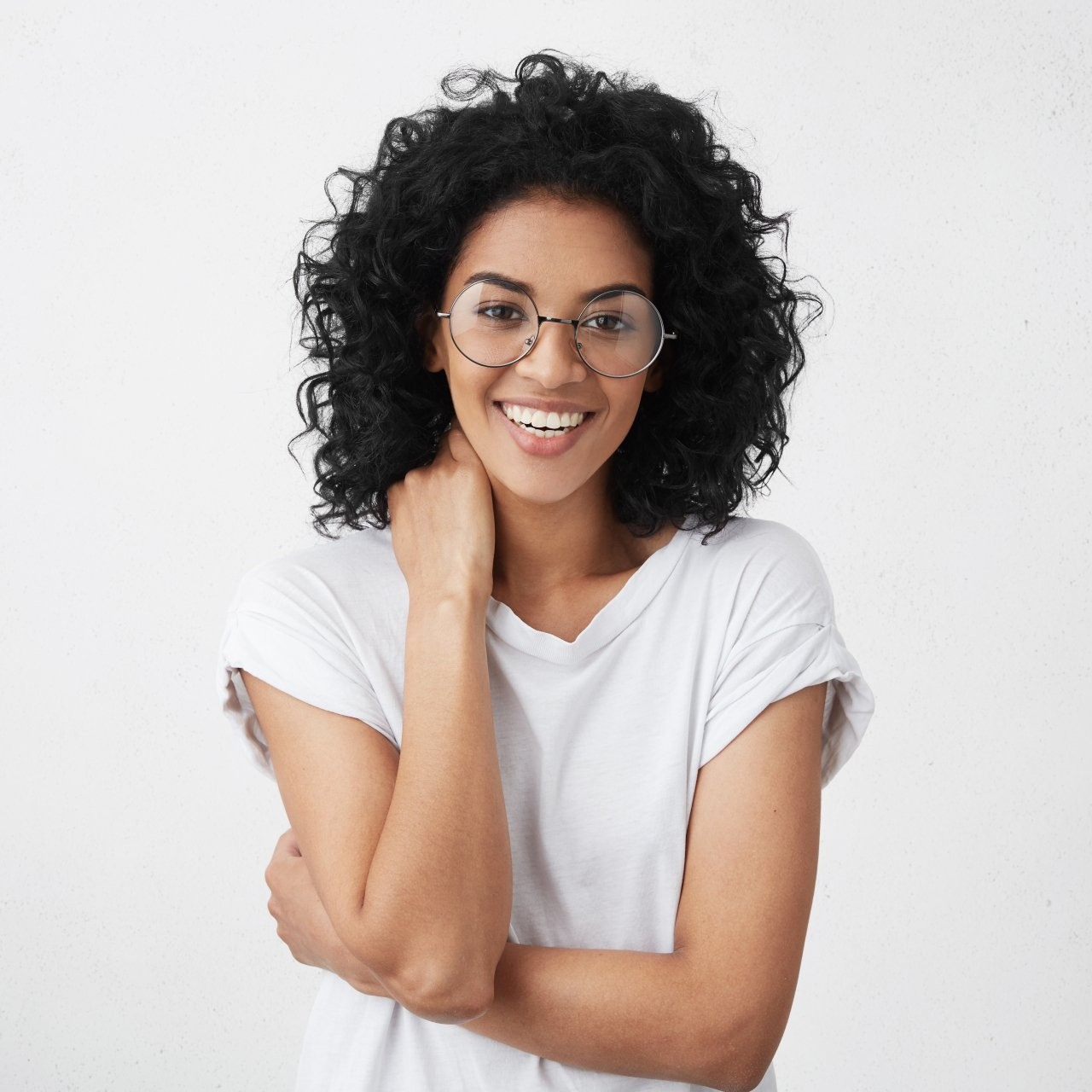 Mujer con cabello negro rizado y gafas sonríe, vistiendo una camisa blanca y con la mano en el cuello.