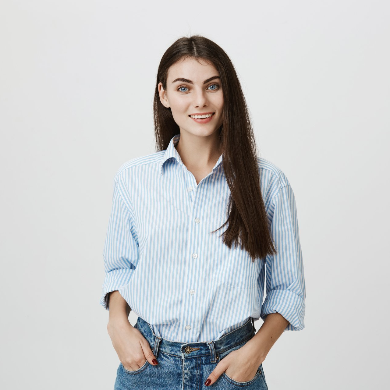 Mujer con cabello largo y castaño, vestida con una camisa de rayas azules y jeans, sonriendo, con las manos en los bolsillos.