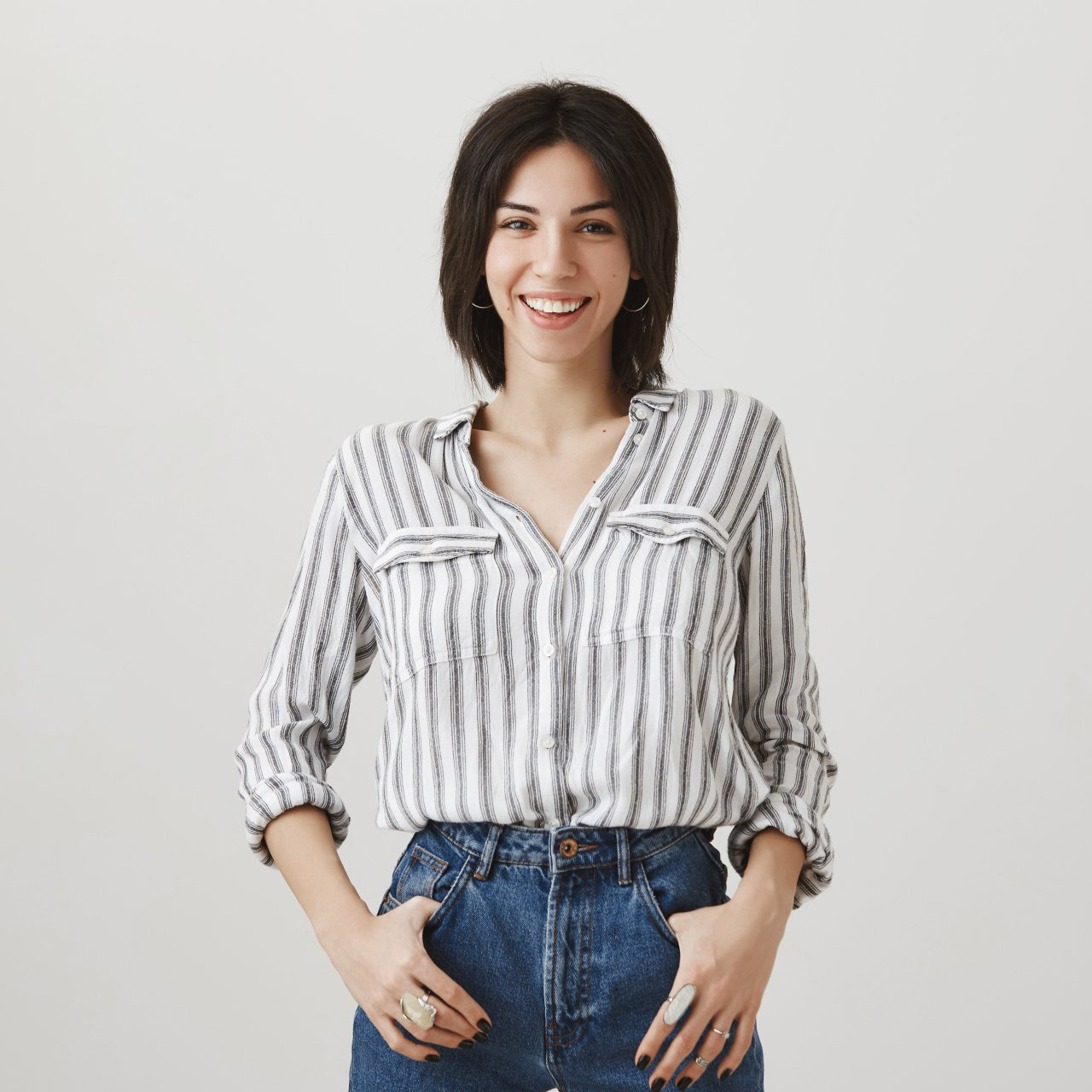 Mujer de cabello oscuro sonríe, viste una camisa a rayas y jeans, con las manos en los bolsillos.