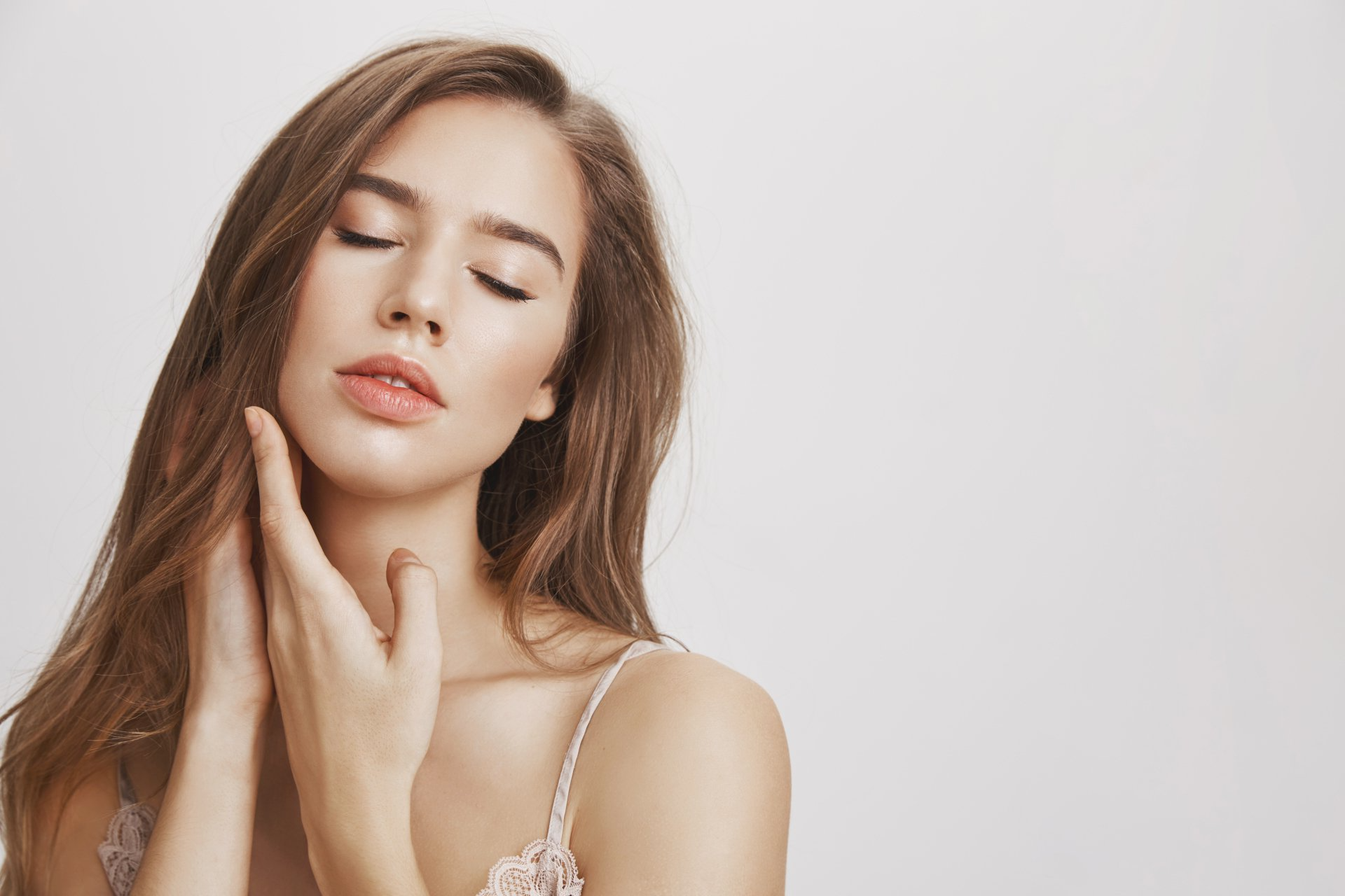 Mujer con los ojos cerrados, tocándose la cara con las manos, piel clara, camisola rosa claro, sobre fondo blanco.