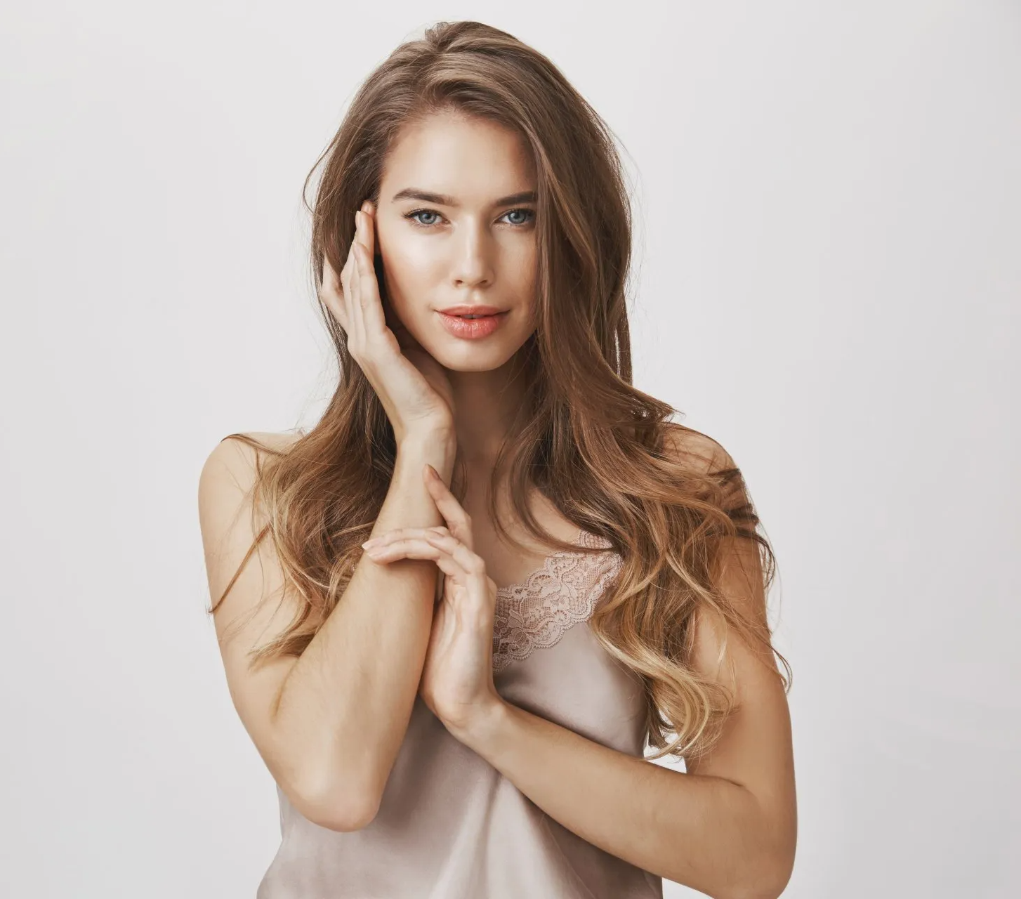 Mujer con cabello largo castaño y una camisola rosa claro, manos cerca de la cara, sonriendo.