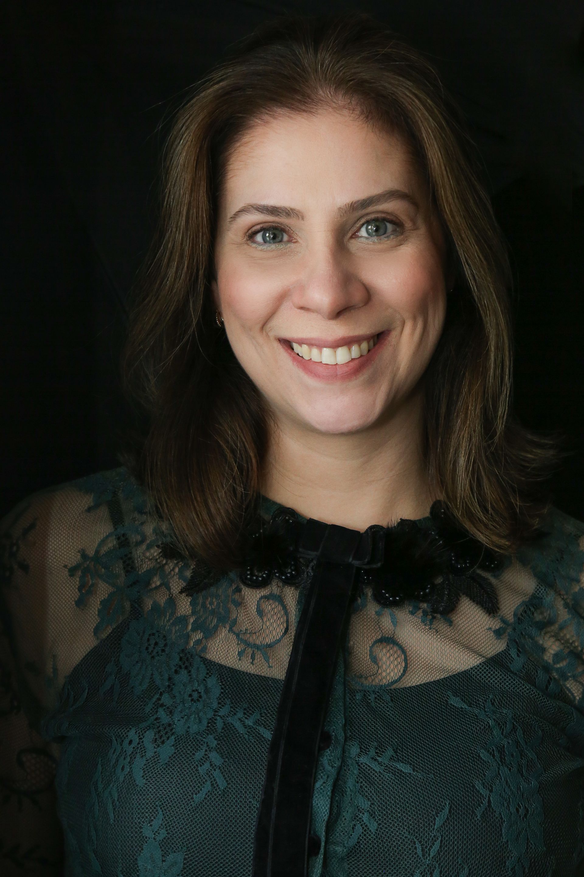 Mulher sorridente, vestindo uma blusa de renda verde-escura com detalhes em preto, contra um fundo escuro.