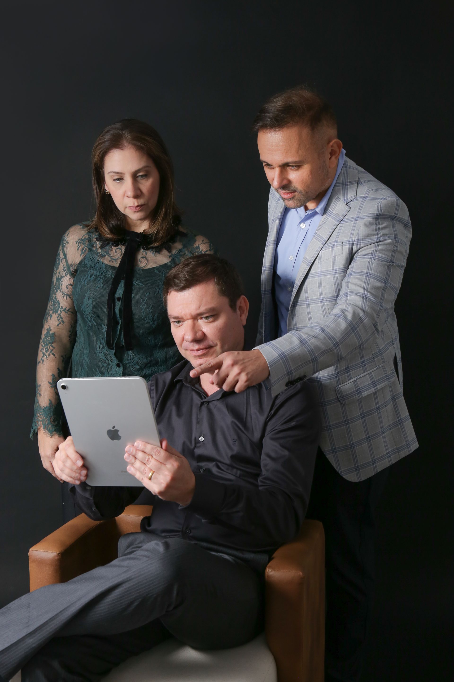 Três pessoas olhando para um tablet contra um fundo escuro. Um homem aponta para a tela enquanto os outros observam.