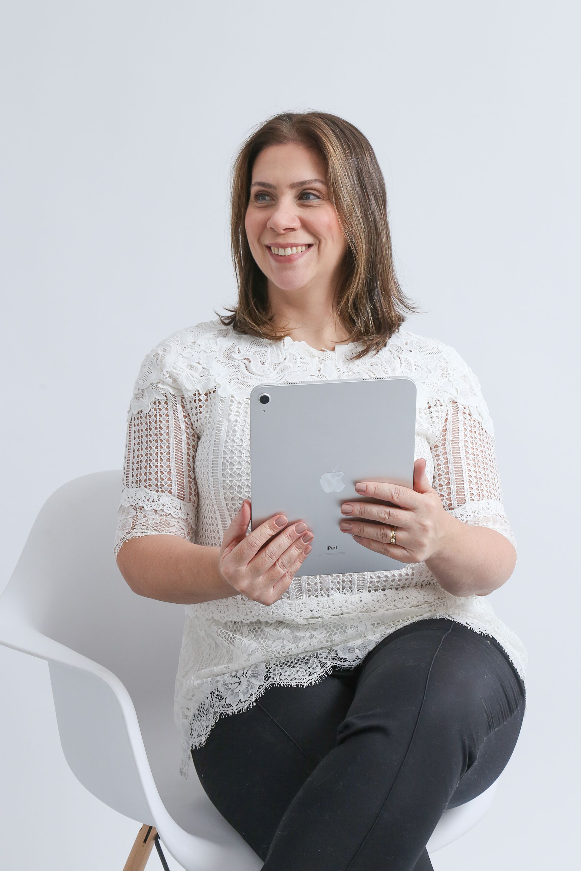 Mulher com blusa de renda branca, sorrindo, sentada em uma cadeira branca contra um fundo branco.