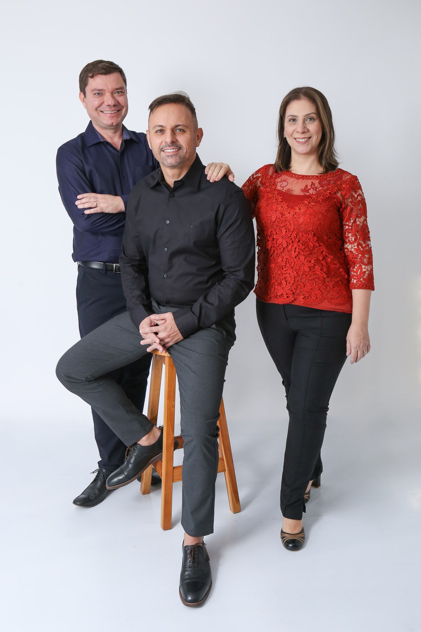 Três pessoas posando: um homem sentado em um banquinho, ladeado por outras duas pessoas. Foto de estúdio contra fundo branco.