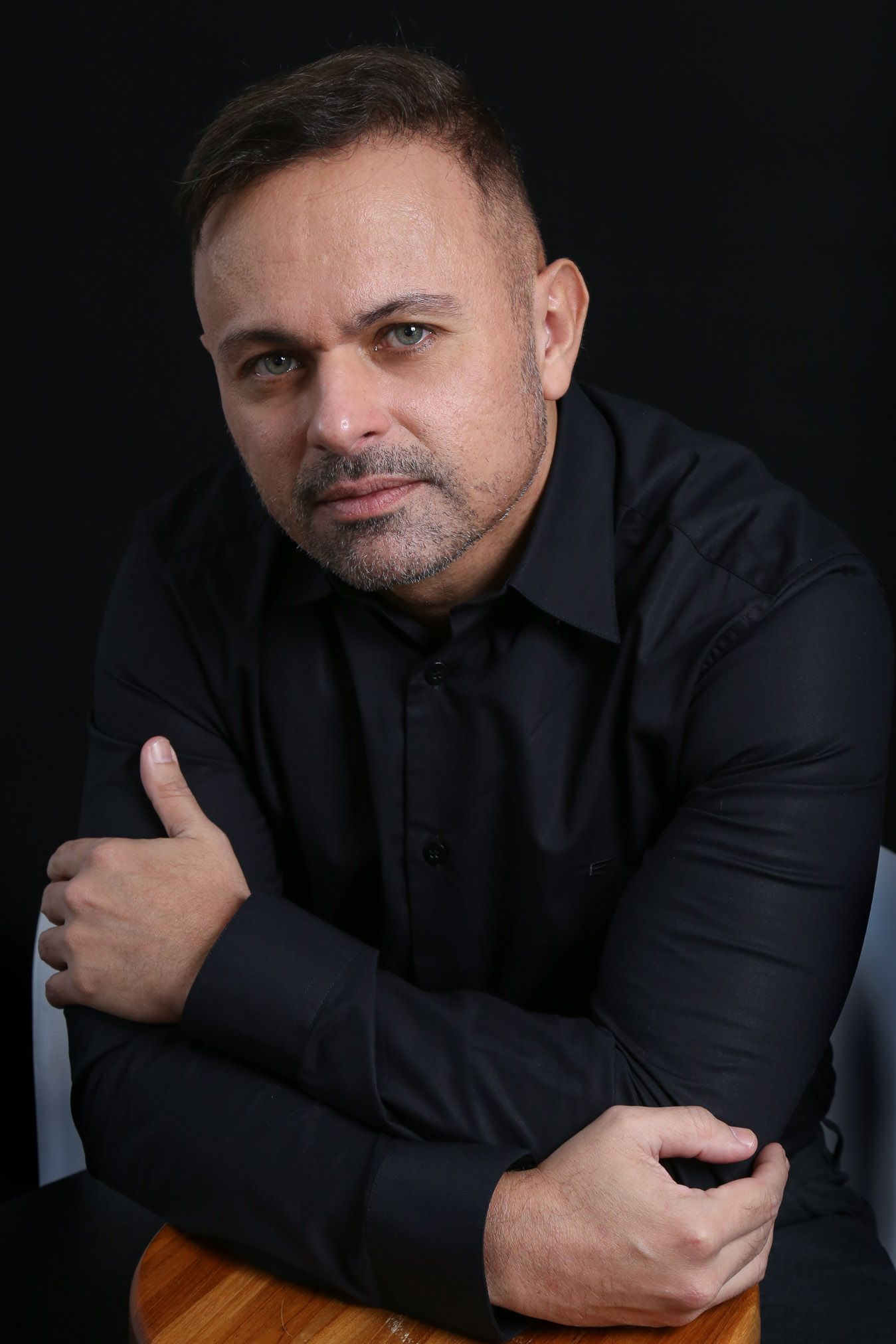 Homem de camisa preta, com os braços cruzados, sentado contra um fundo preto, olhando para a câmera.