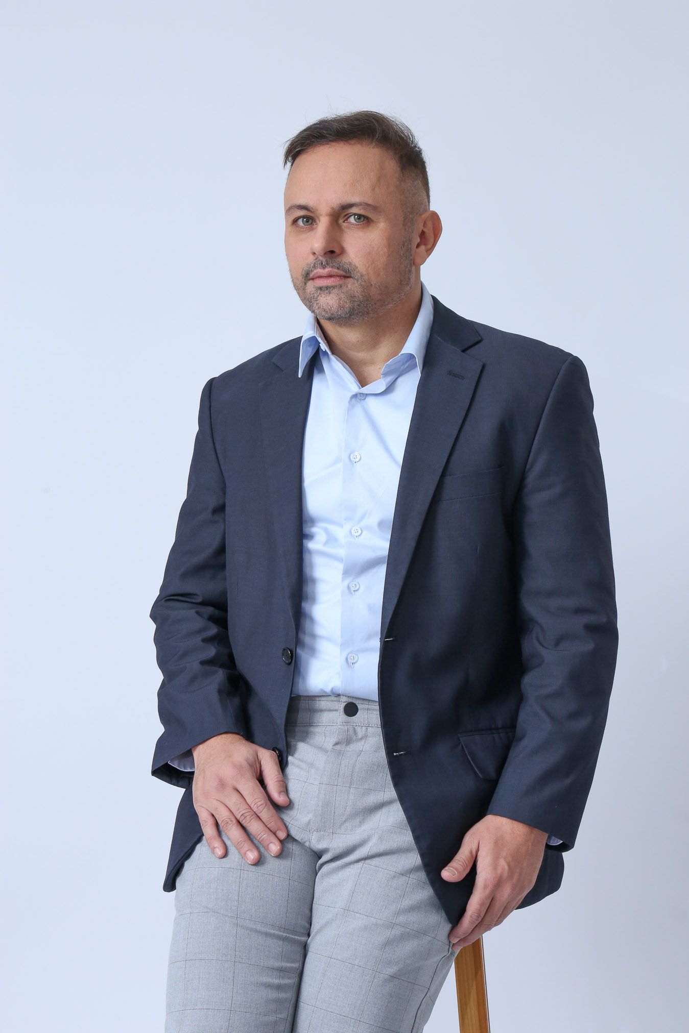 Homem de blazer azul, camisa azul clara e calça cinza, encostado em um banquinho, olhando para a câmera.