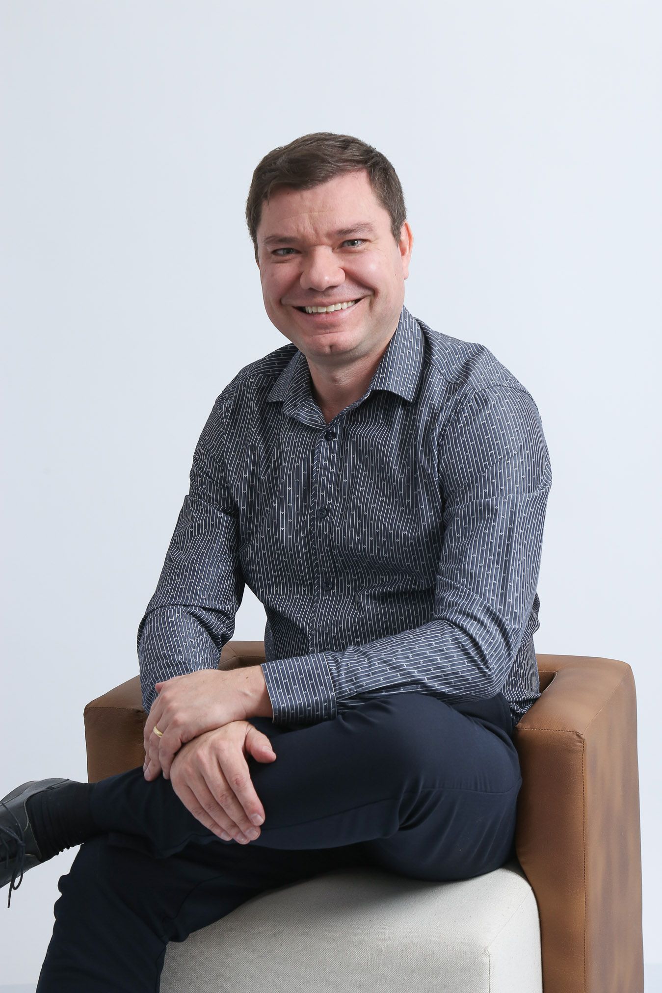 Homem sorrindo, sentado em uma cadeira marrom, com as pernas cruzadas.