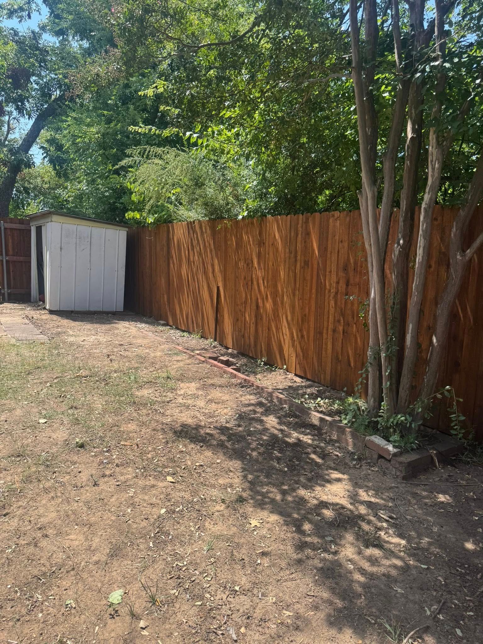 Brown wooden fence next to a dirt yard with a small shed and trees in the background.