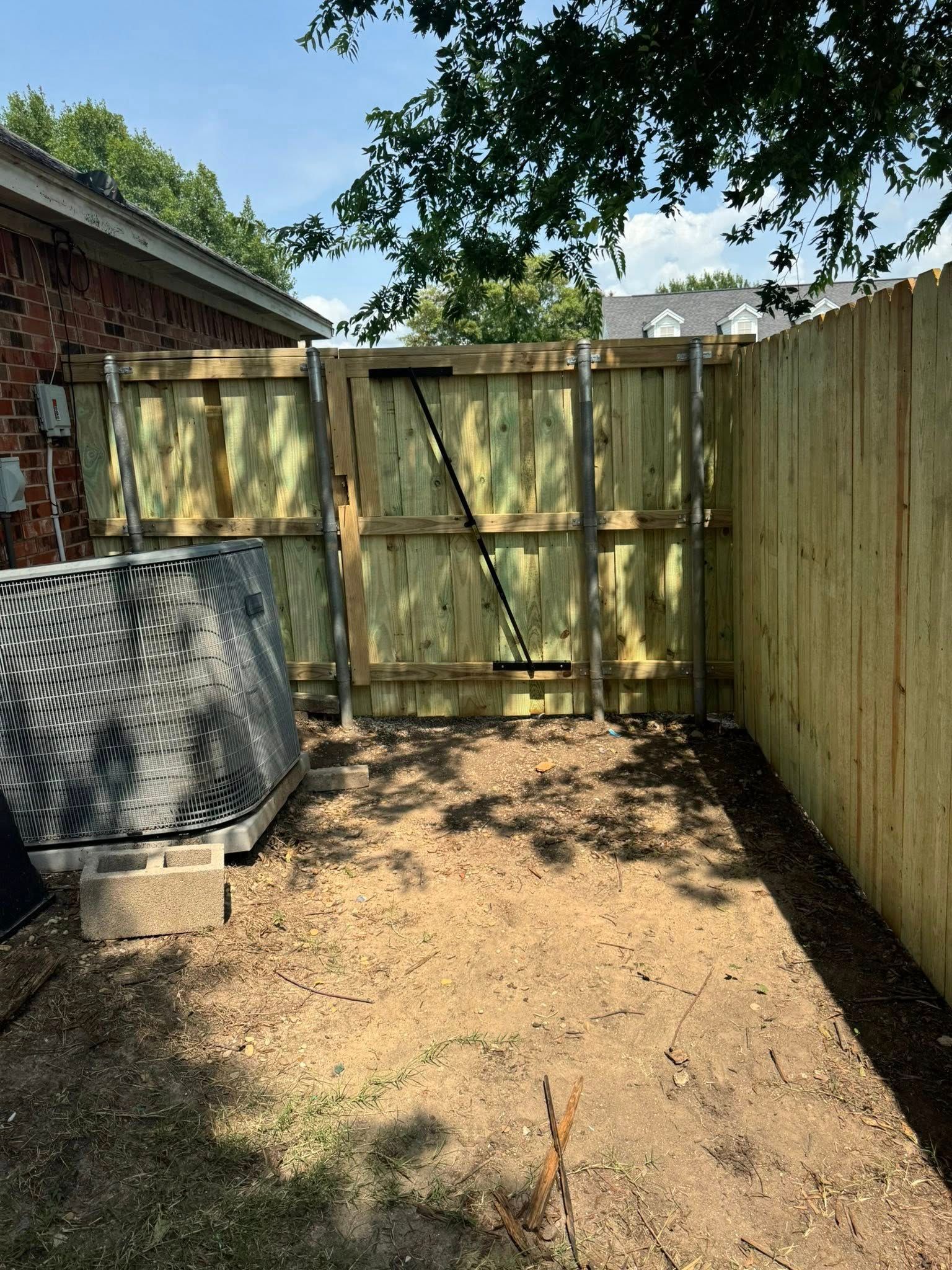A small, dirt-floored backyard enclosed by a wooden fence and gate, with an air conditioning unit.