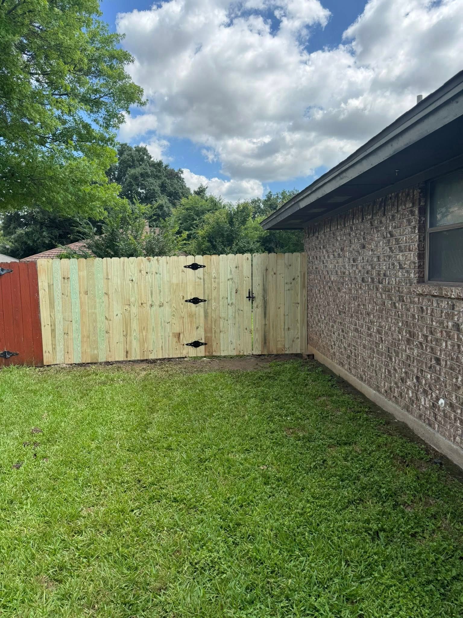 Wooden fence with black hardware adjoins a textured wall; green grass and a cloudy sky.