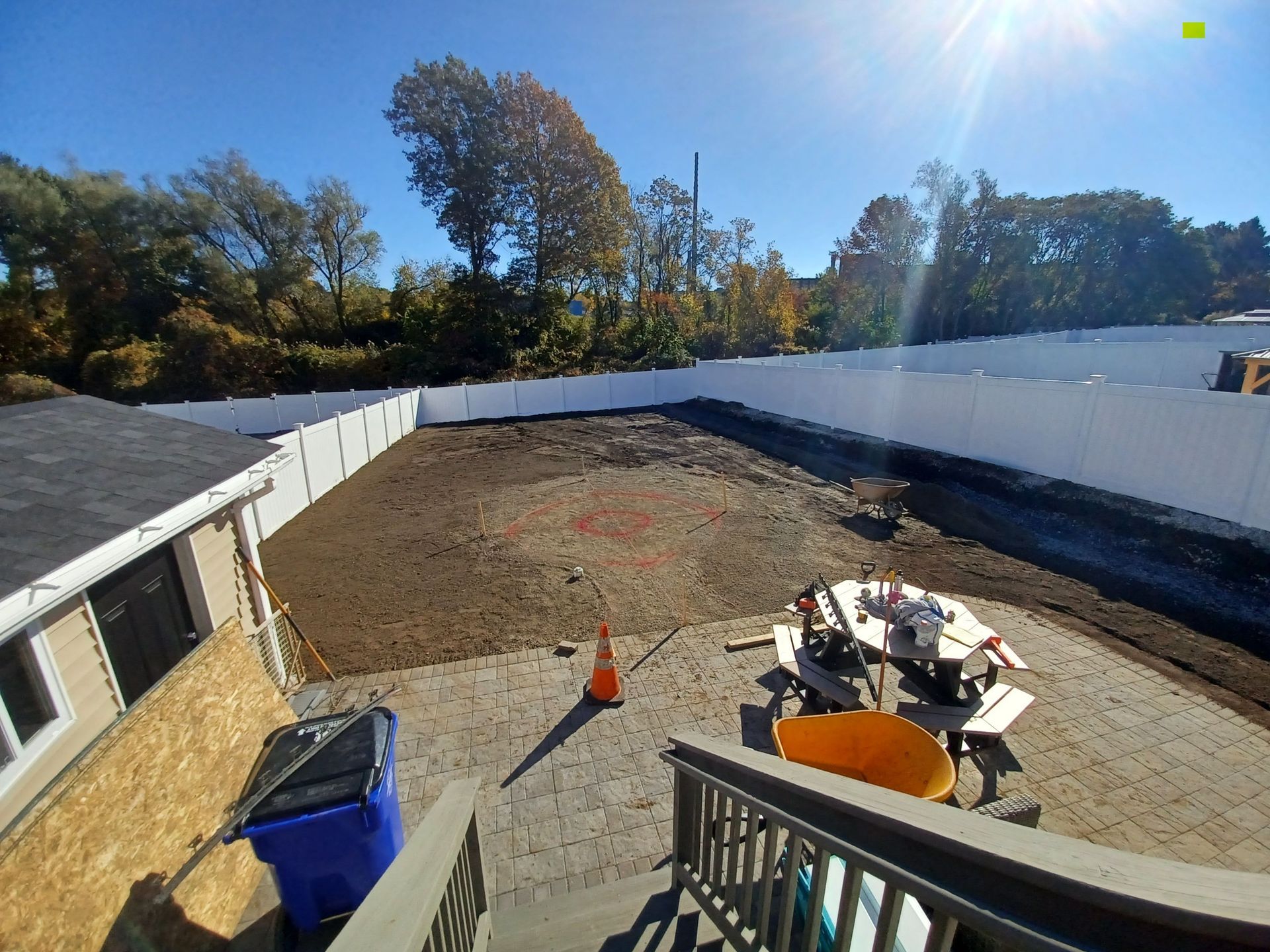A backyard view with a patio, a dirt patch marked with orange spray paint, and a white privacy fence against a tree line.