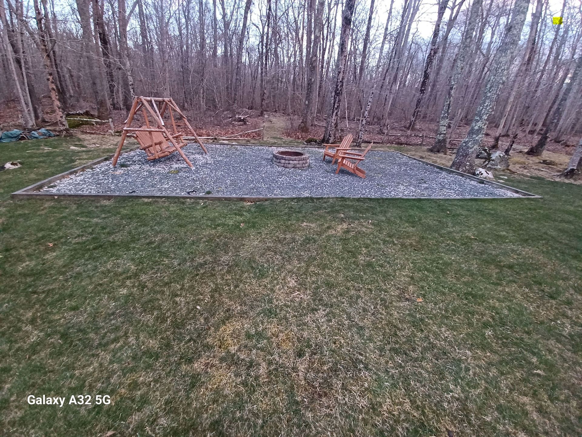 A wooden swing set and fire pit sit on a gravel patio in a grassy backyard, surrounded by a dense forest of bare trees.