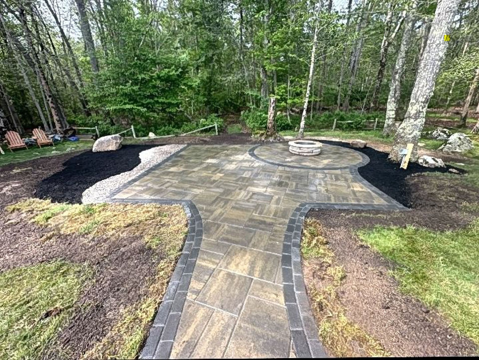A stone paver patio and walkway lead to a circular fire pit in a wooded backyard, framed by black mulch and lawn.