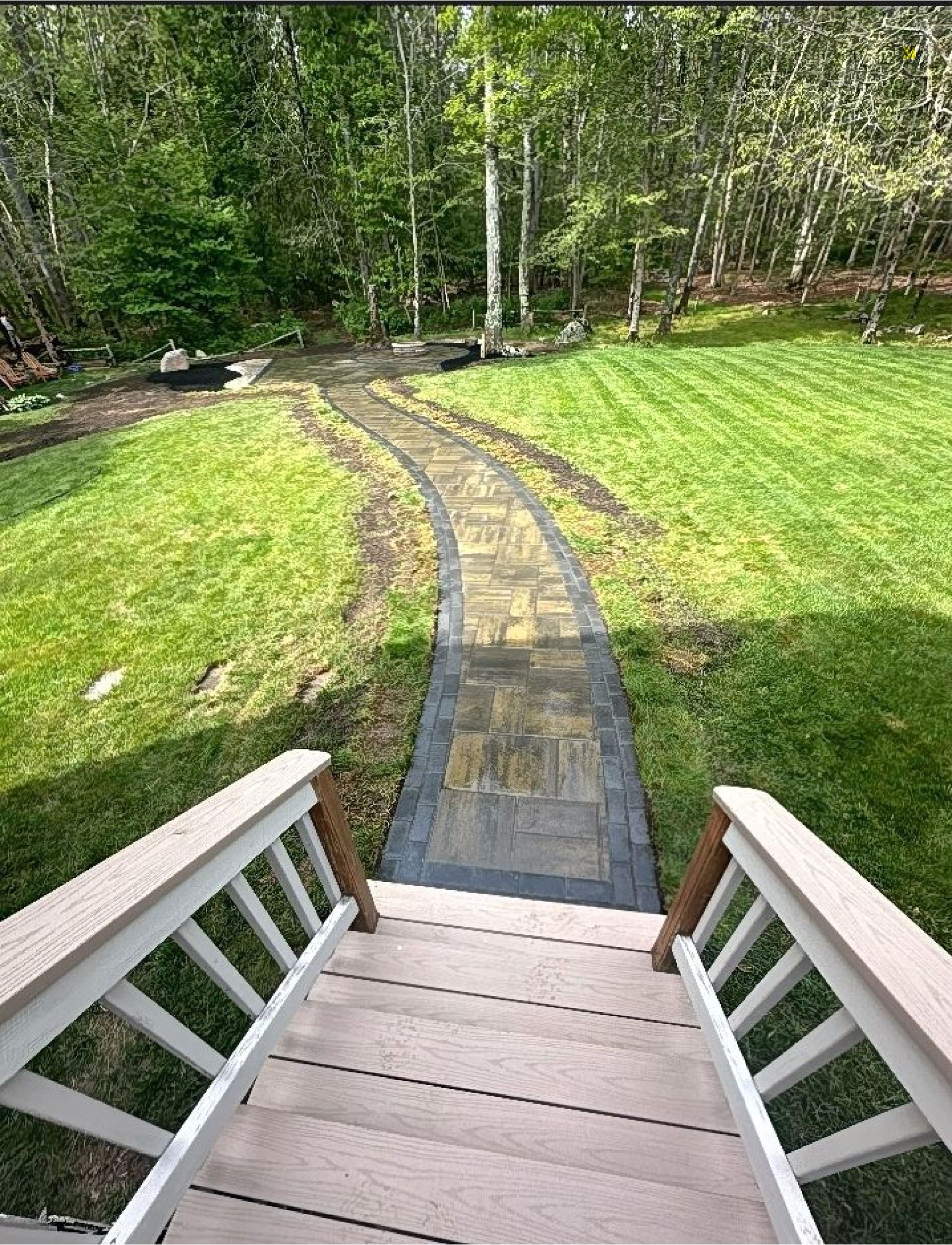 A view from the top of wooden porch steps looking down onto a stone paver path leading through a grassy yard to trees.