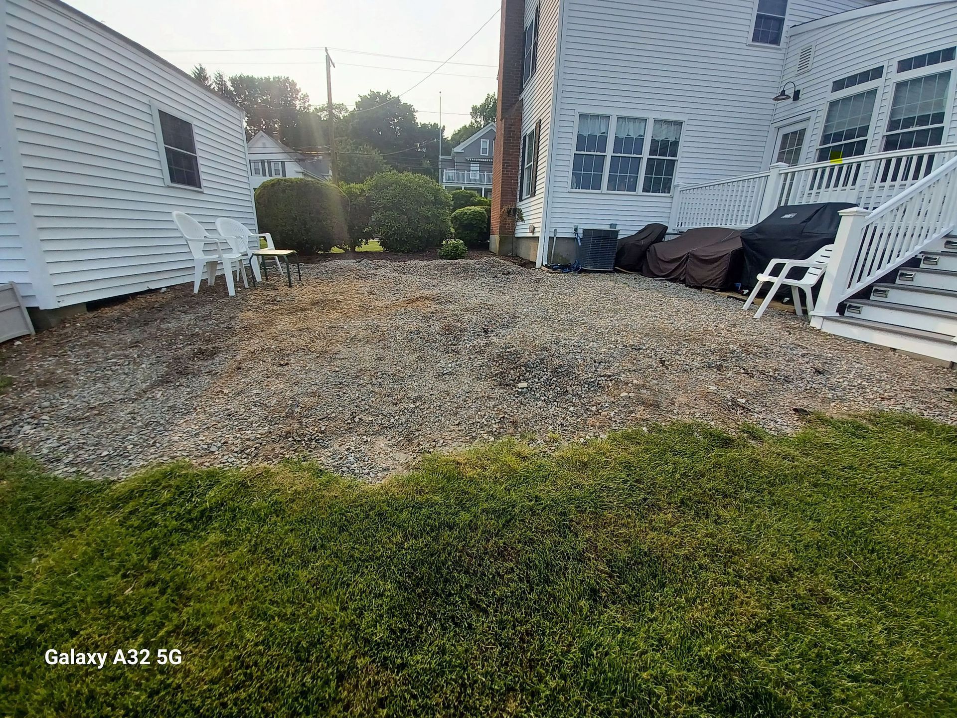 A gravel backyard patio area sits between a white garage and a multi-story white house with a deck and stairs.