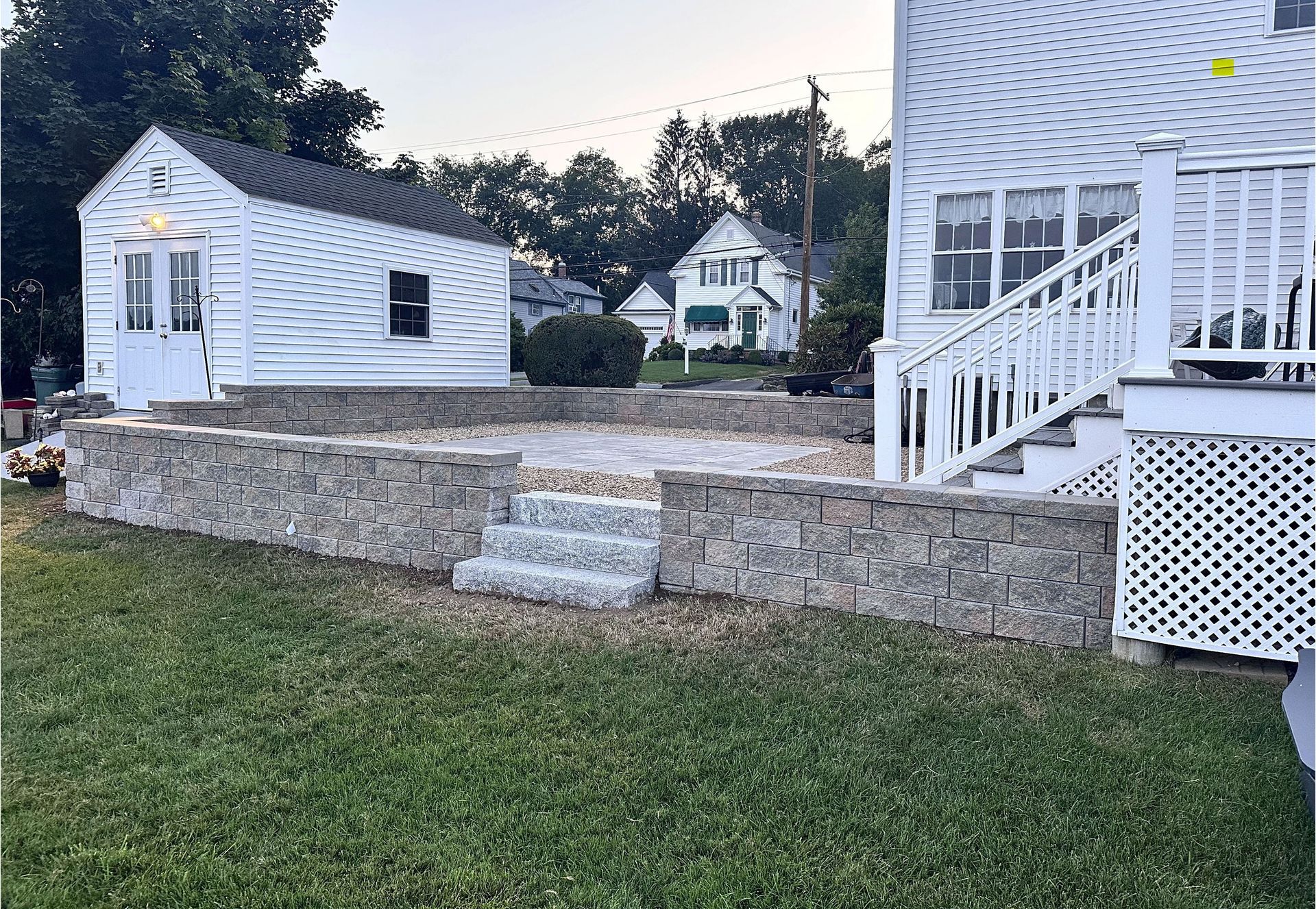 A stone retaining wall surrounds a patio with steps in a grassy backyard, situated between a house and a small shed.