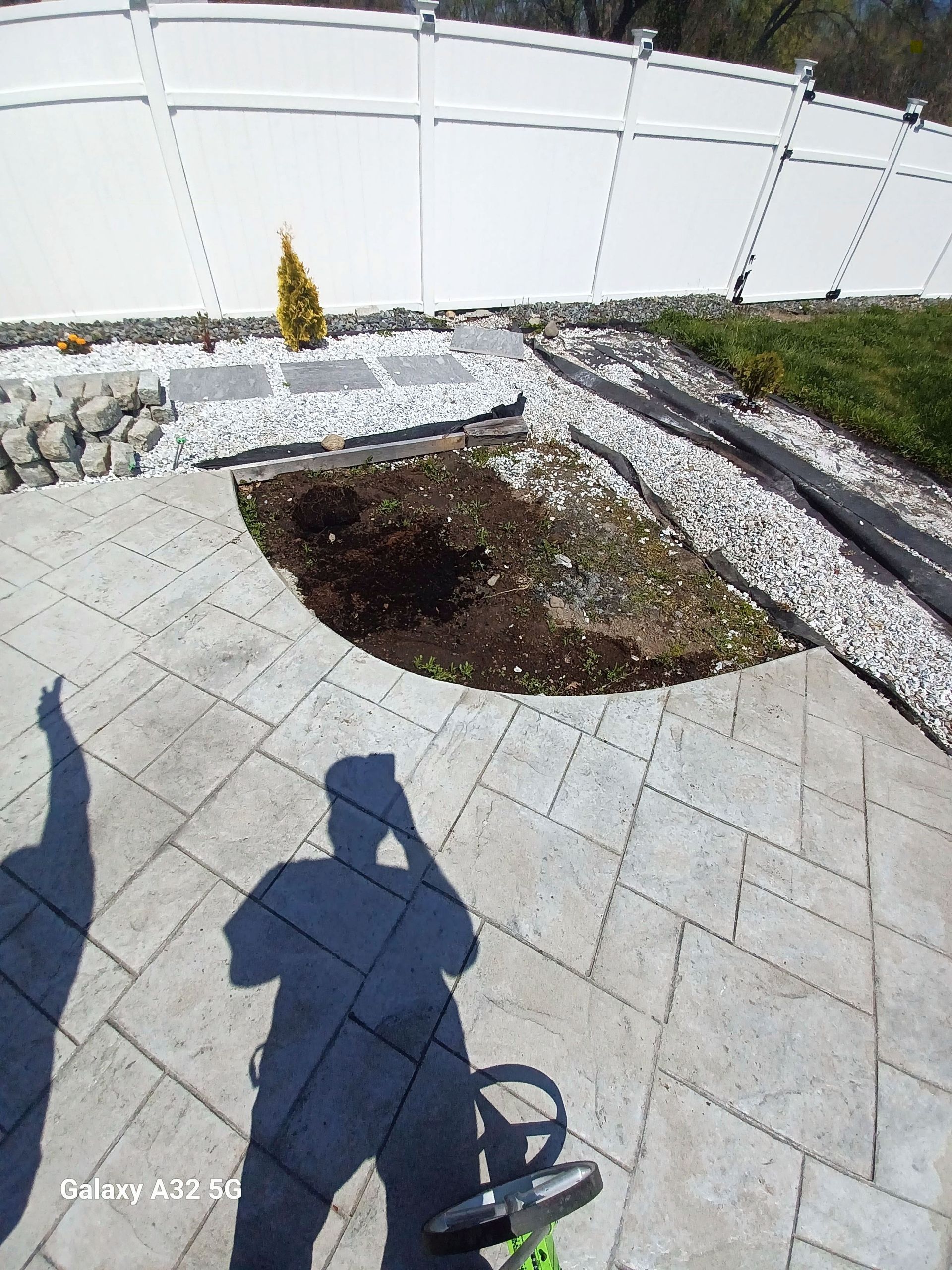 A paved patio featuring a curved cutout in the center filled with soil, adjacent to a decorative white rock border.