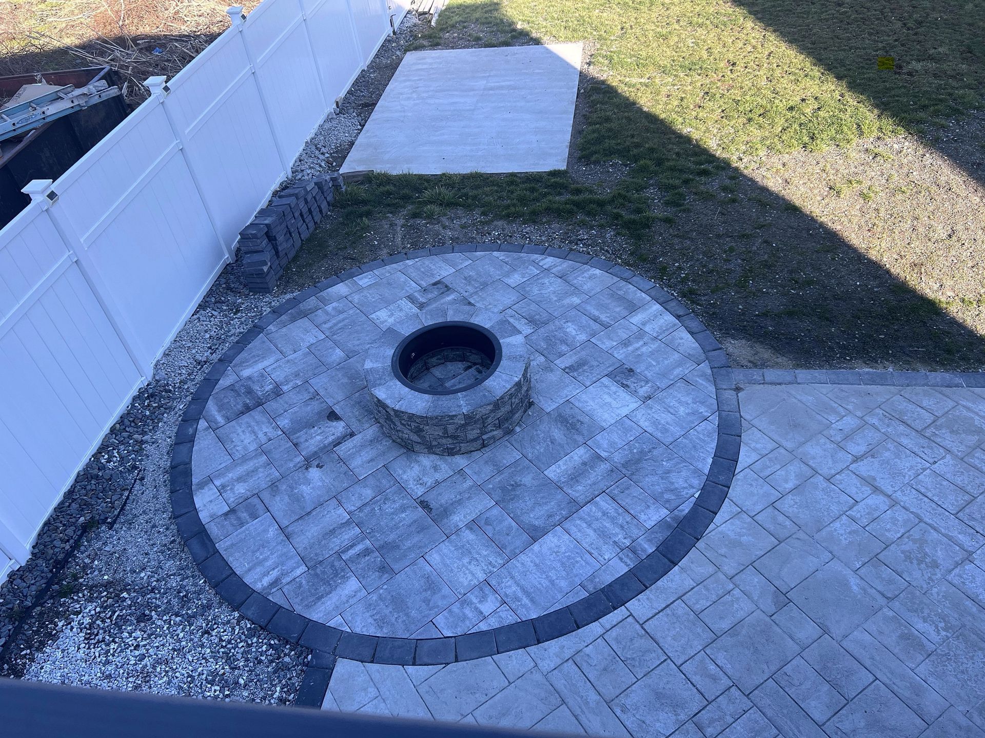 A circular stone fire pit patio area with a white vinyl fence and rectangular paver walkway in a backyard setting.