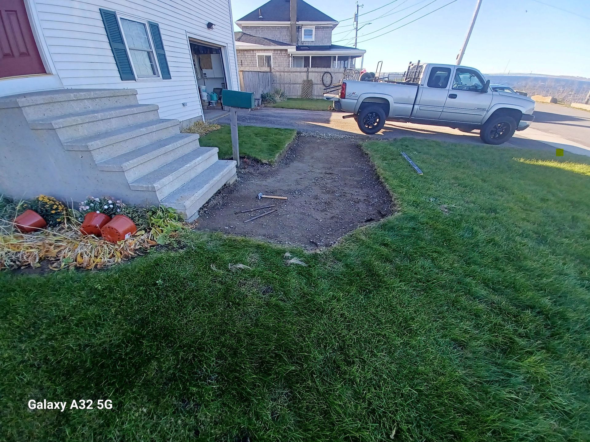 A front yard with concrete steps leading to a house, an adjacent bare dirt garden bed, and a pickup truck on the street.