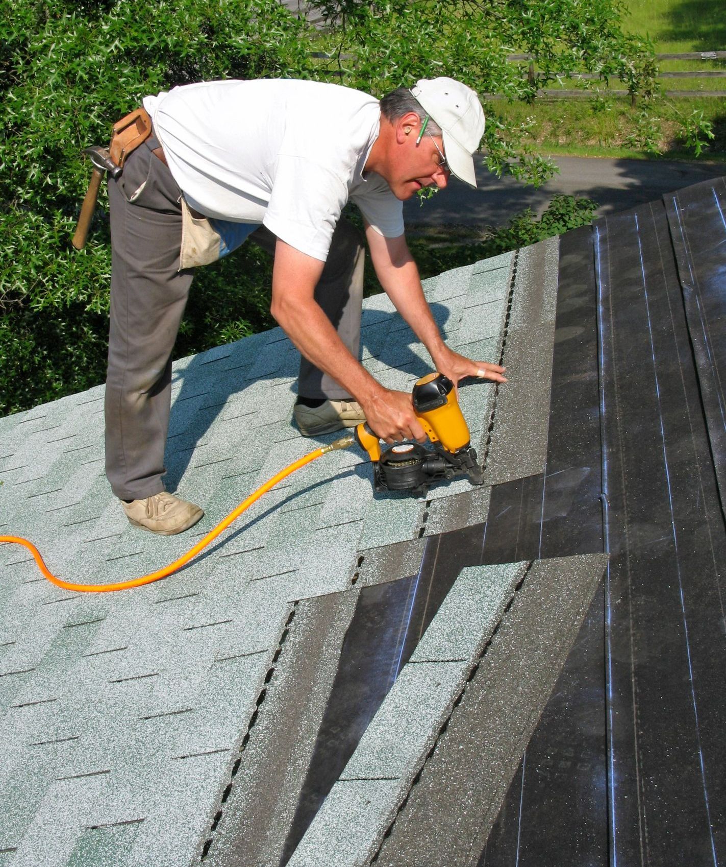A worker is installing asphalt shingles on a roof using a nail gun.