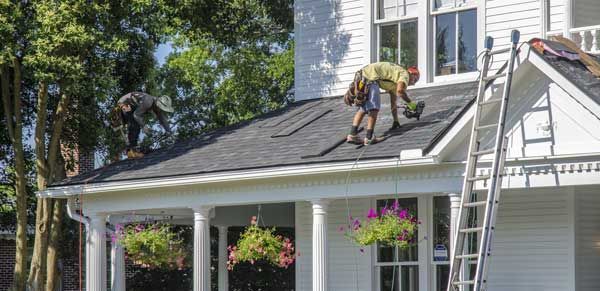 Two workers on a roof, installing shingles. A ladder leans against the house.
