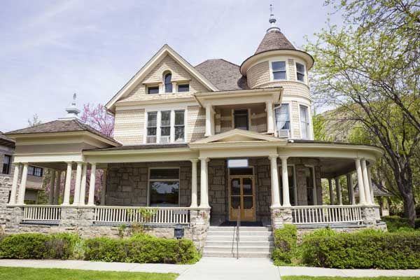 Two-story stone house with wrap-around porch, turret, and decorative roof, fronted by a lawn.