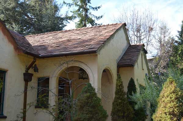 Tan stucco house with a brown tile roof, arched entryway, and evergreen shrubs.
